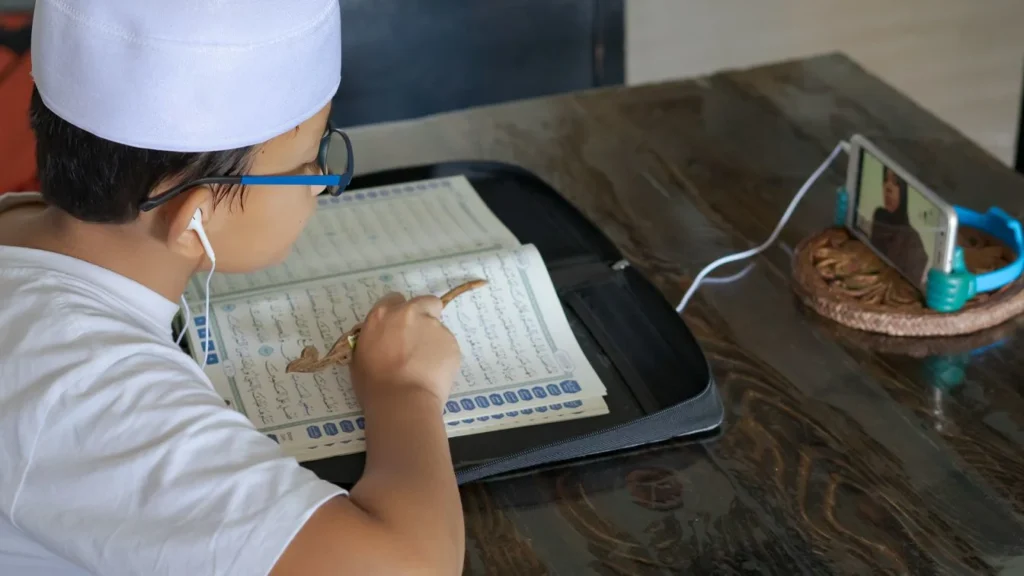 Child attending an online Quran learning class while reading the Quran and following a teacher on a smartphone.